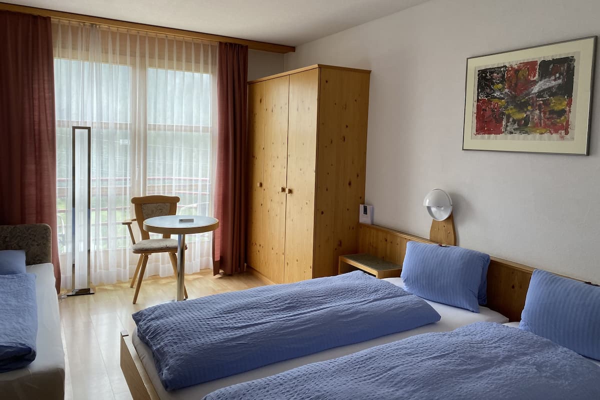 Habitación de hotel con dos camas, un armario de madera, una mesita pequeña, una silla y un ventanal con cortinas traslúcidas.