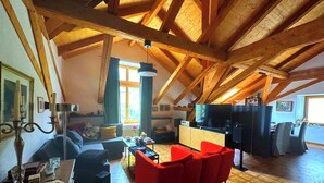 A spacious loft with high ceilings, exposed wooden beams, a cozy seating area with red chairs, a television, and a dining area.