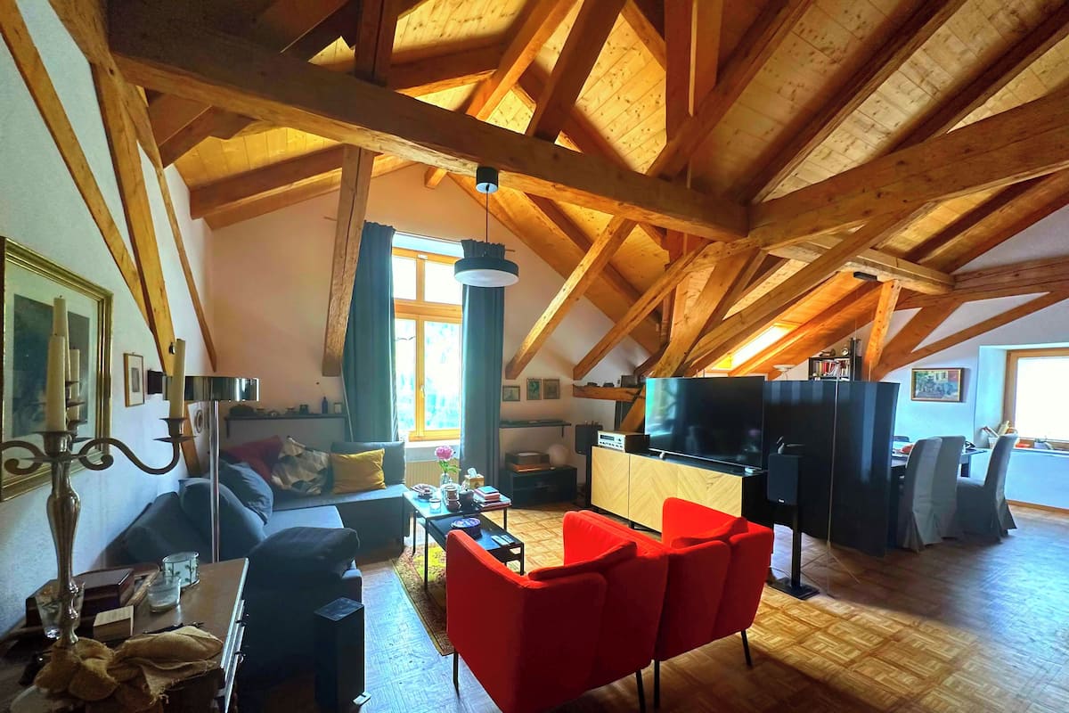 Un loft espacioso con techos altos, vigas de madera a la vista, una zona de estar acogedora con sillas rojas, un televisor y un comedor.