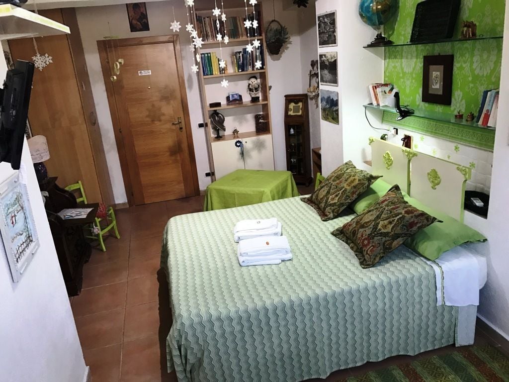 Una camera da letto con una coperta verde, cuscini e un letto rifatto con cura. Si nota una porta in legno, una libreria e un tavolino con tovaglia verde.