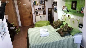 Una camera da letto con una coperta verde, cuscini e un letto rifatto con cura. Si nota una porta in legno, una libreria e un tavolino con tovaglia verde.
