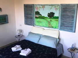 Un dormitorio con cama, dos almohadas, mesita de noche con lámpara, cuadro en la pared y ventana con vista a un campo y árboles.