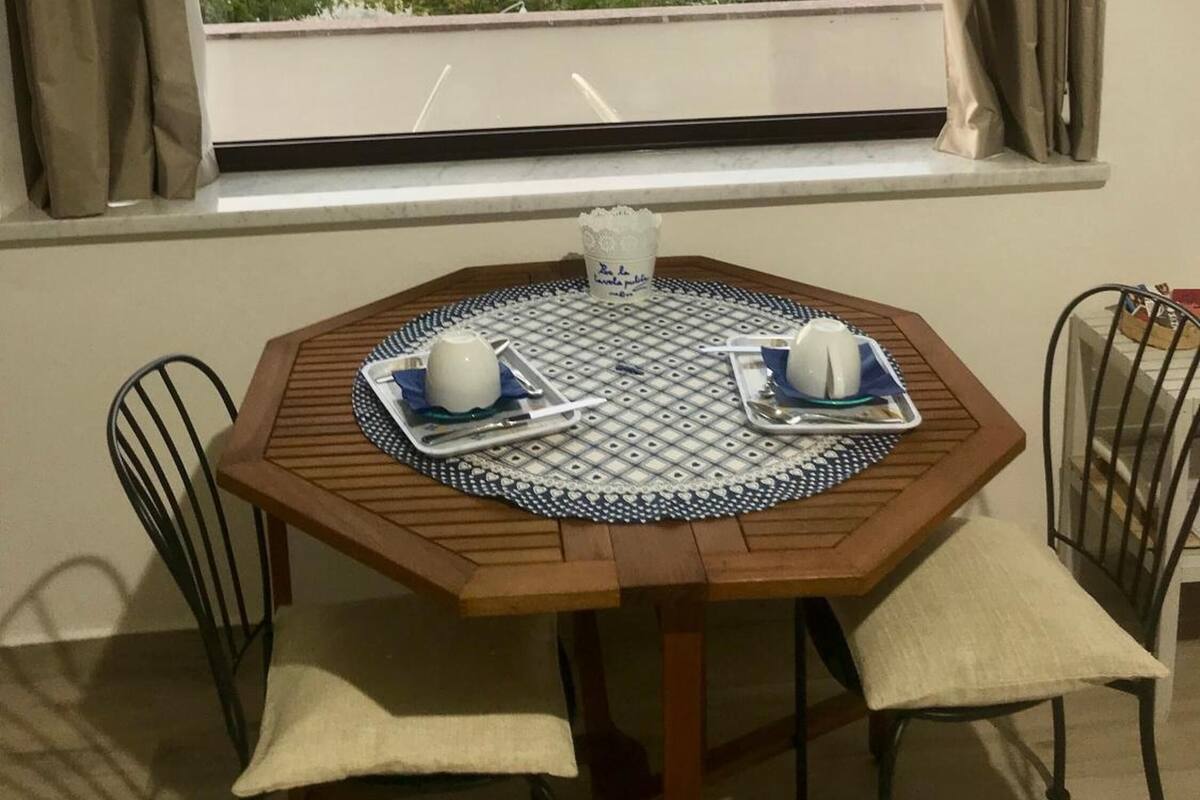 A dining area with a table set for two, a window with curtains, and a view of the sea and greenery.