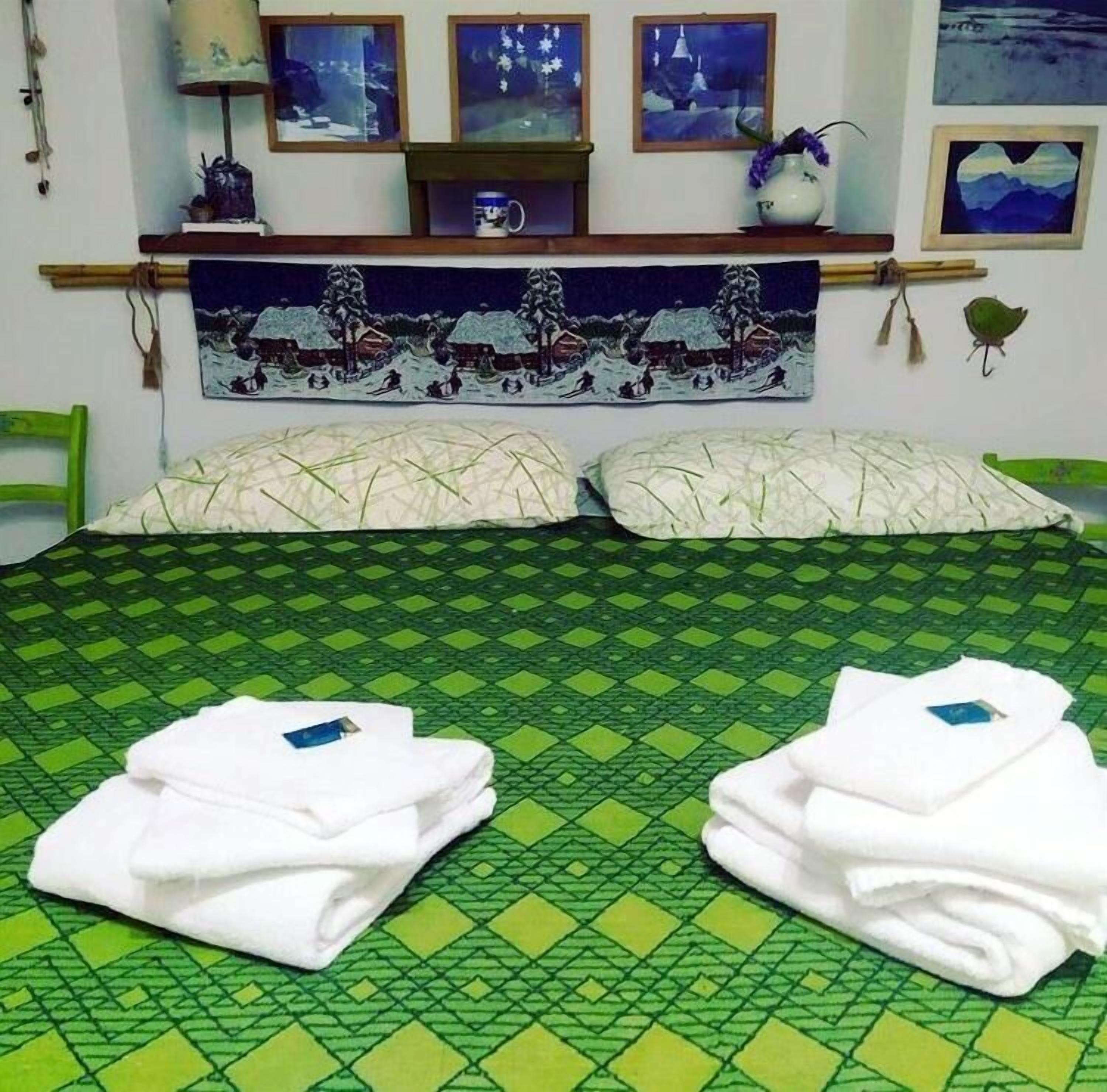 A bed with a green patterned bedspread, two pillows, and two folded white towels on a green and yellow checkered floor.