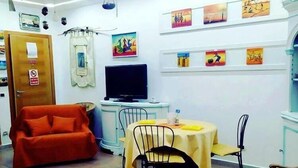Una habitación pequeña y luminosa con un sofá, una mesa con sillas, un televisor y obras de arte en las paredes.