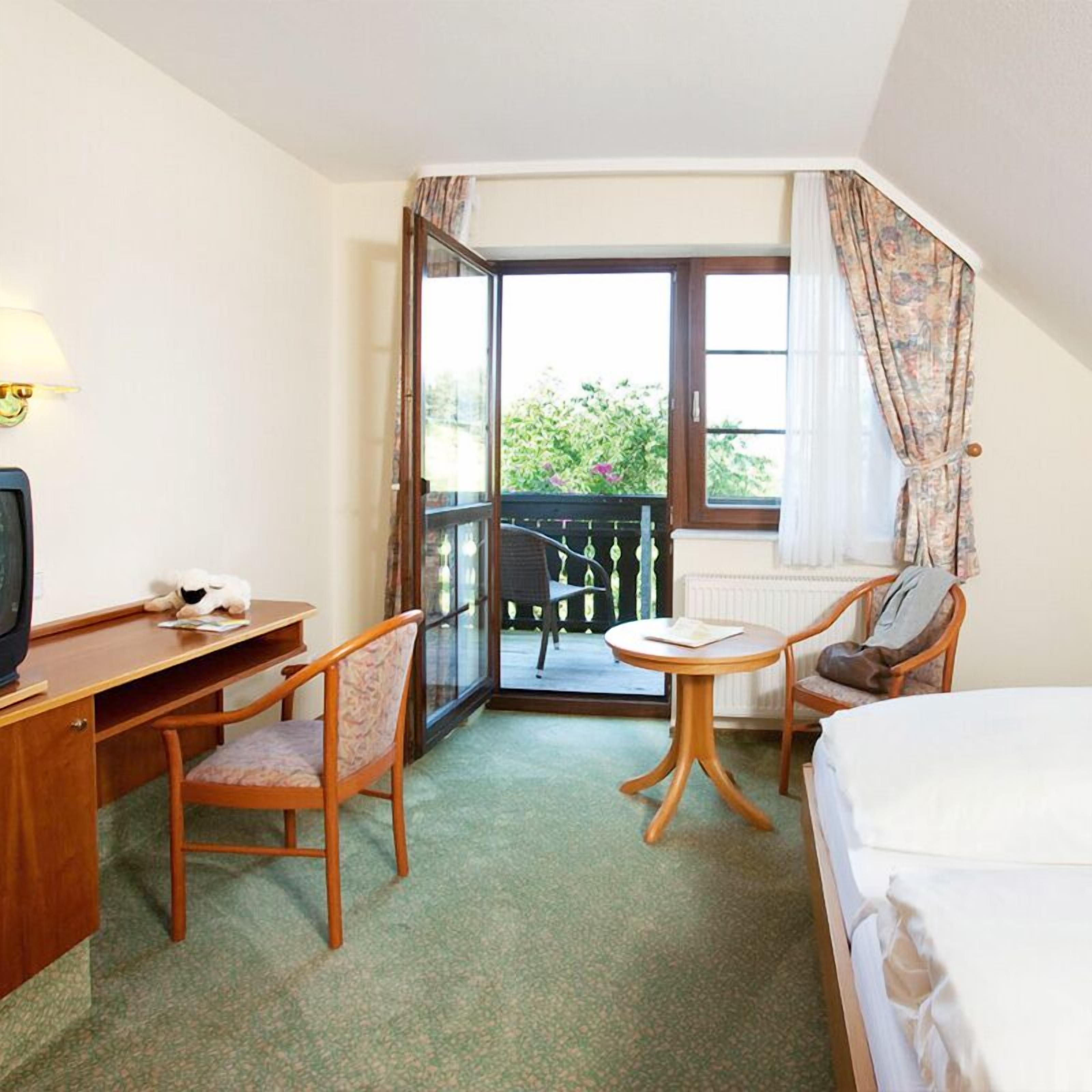 Habitación de hotel con cama, escritorio y silla, una mesita con otra silla, y un balcón con vistas a zonas verdes.
