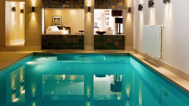 Una piscina interior moderna con suelo y paredes de azulejos, una zona de recepción con mostrador y asientos, y un techo con un diseño de ventilador único.