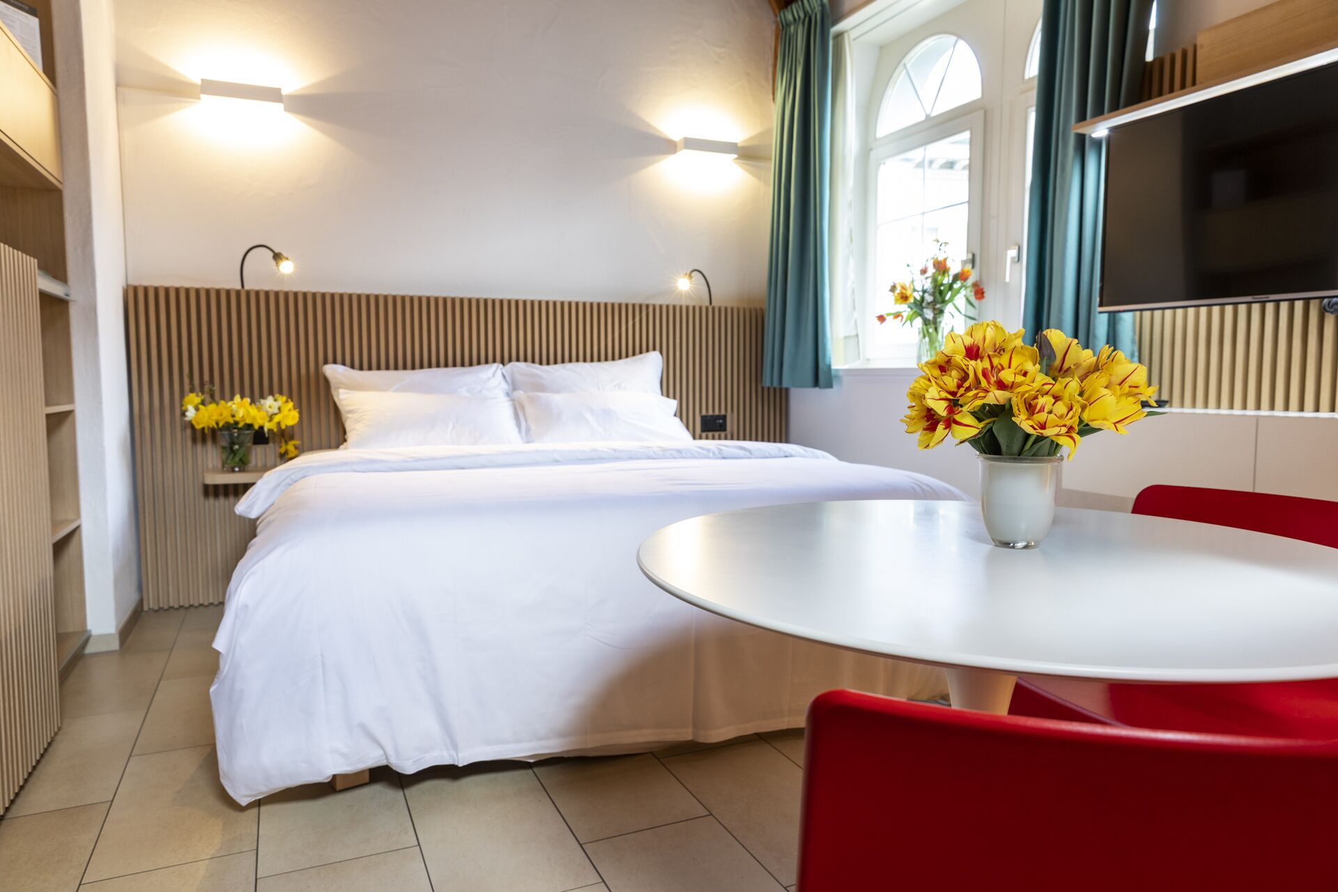 Een hotelkamer met een bed, een nachtkastje met bloemen, een ronde tafel met stoelen en een televisie aan de muur.