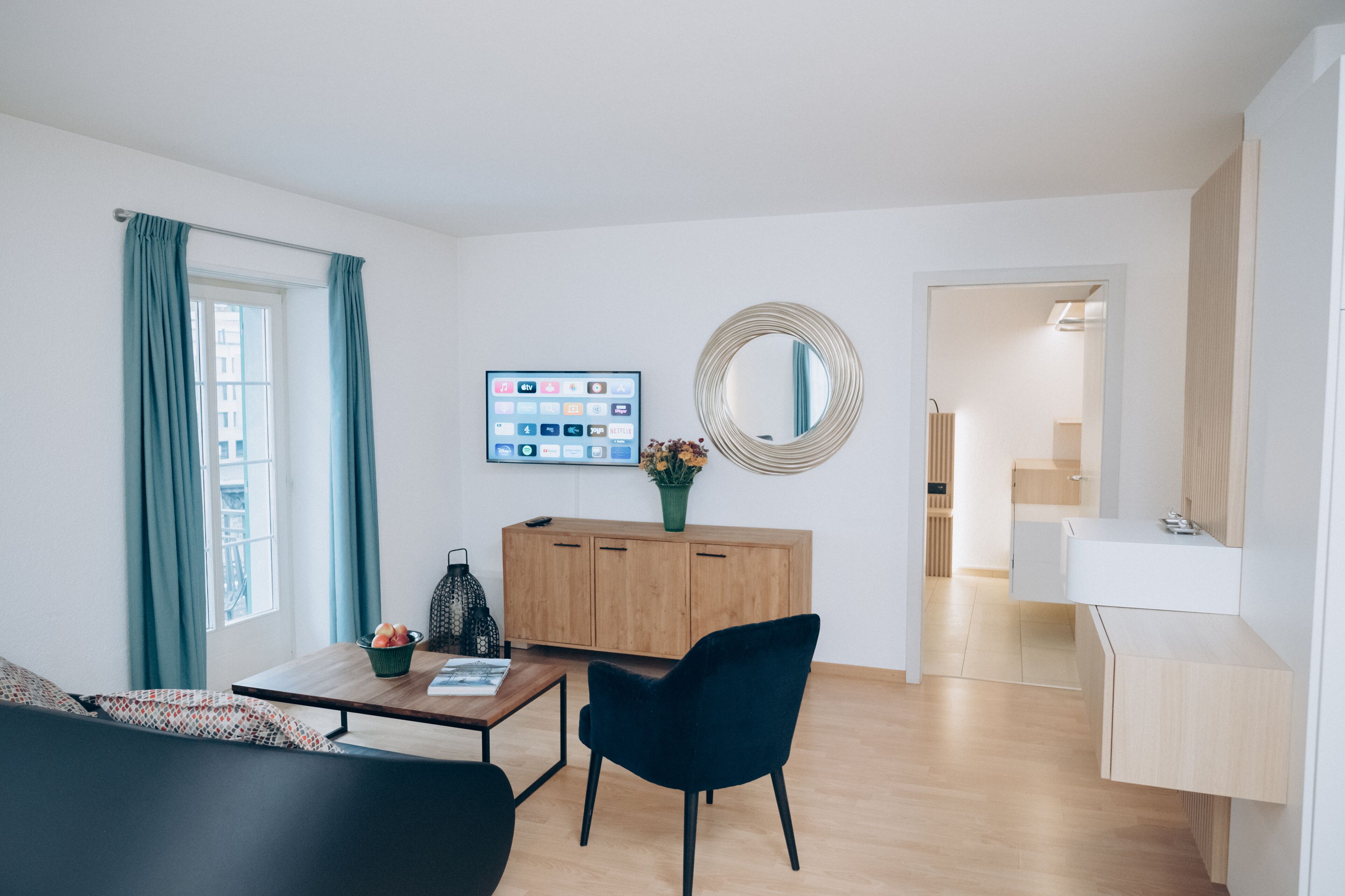 Een moderne woonkamer met een houten kast, een zwarte stoel, een salontafel met een fruitschaal en een aan de muur bevestigde televisie.