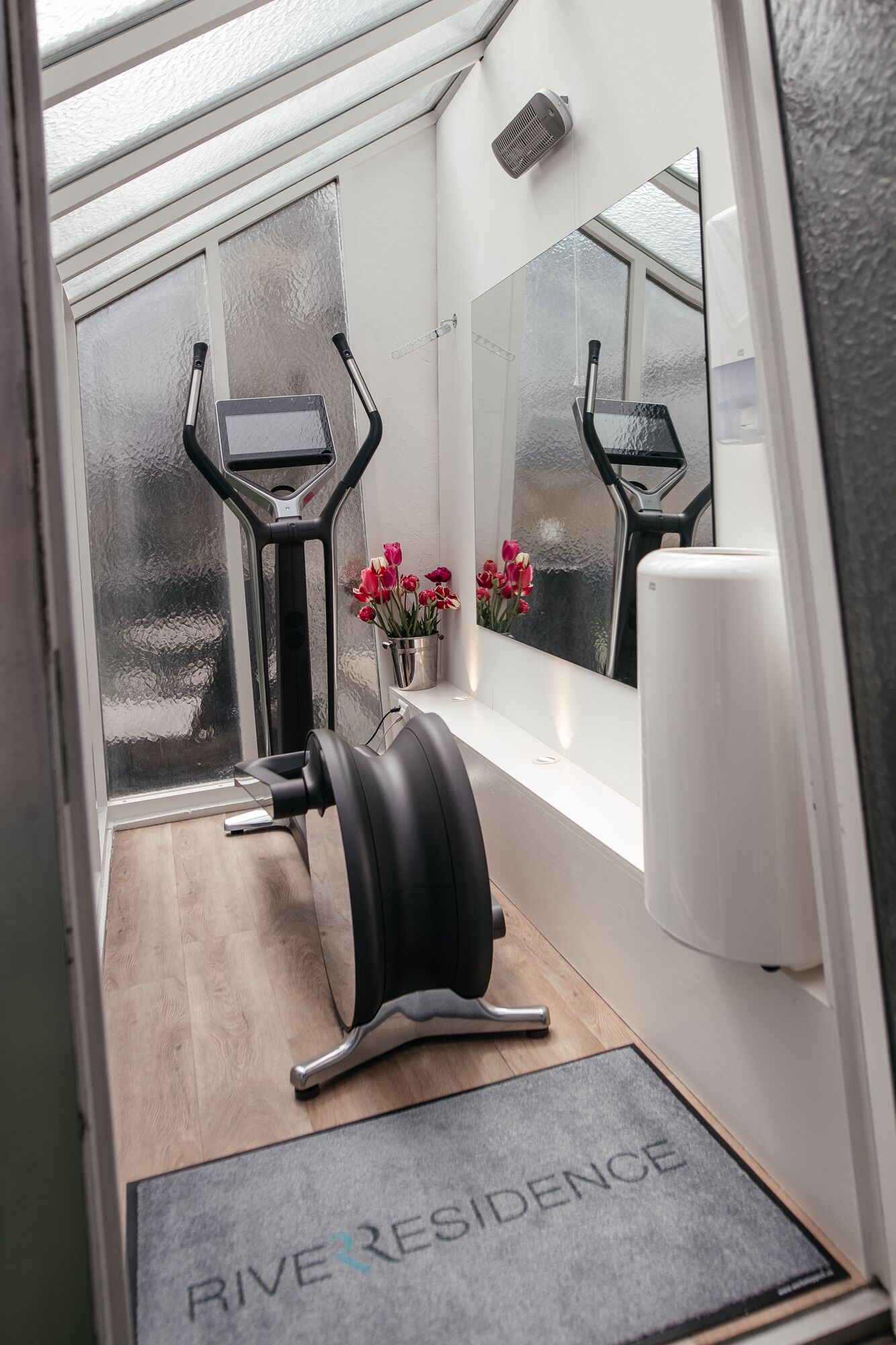 A compact gym with a treadmill, a stationary bike, and a weight bench. A small potted plant with red flowers is on a windowsill, and a mirror reflects the room.