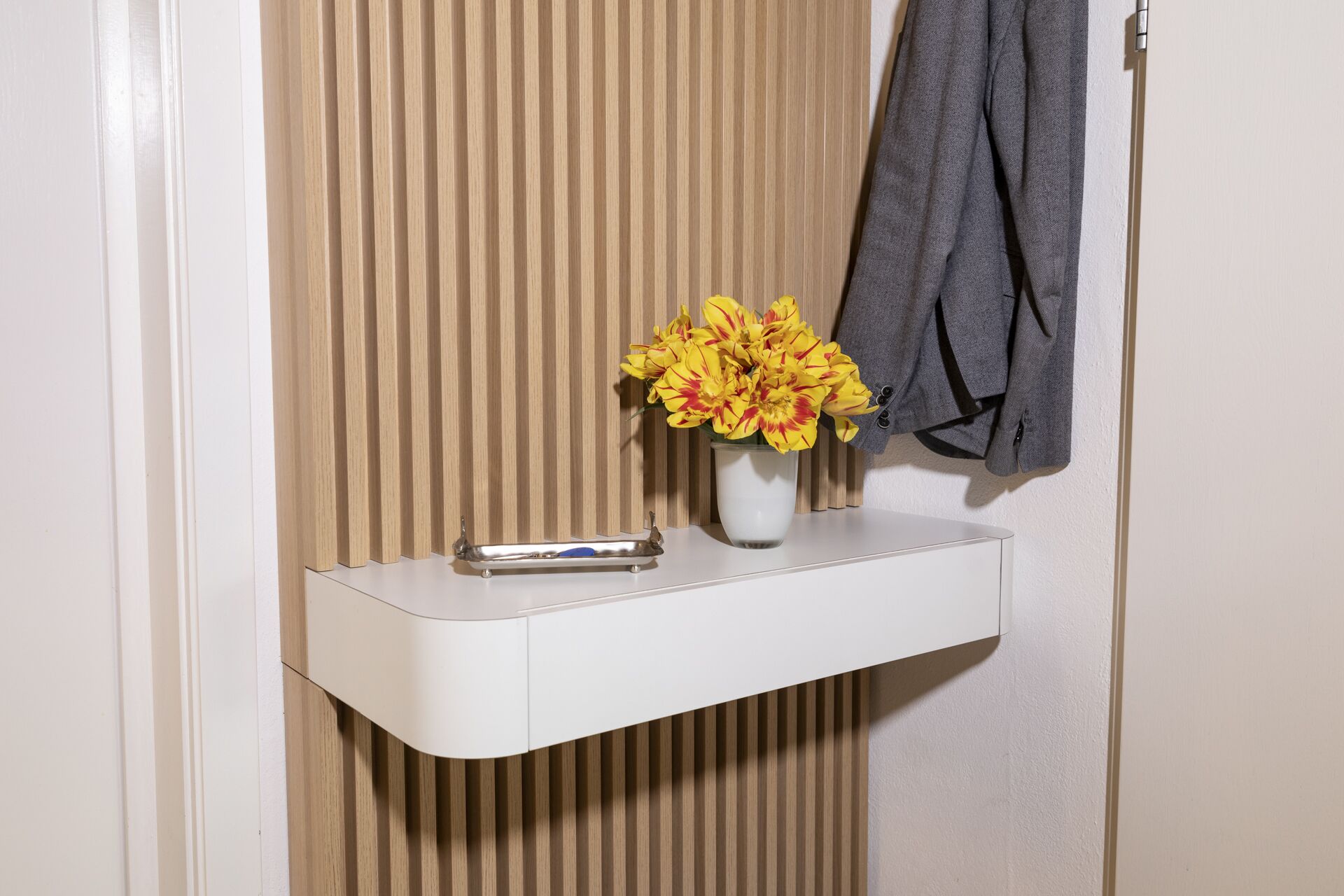 Ein weißer Nachttisch mit einer Vase gelber Blumen, einer metallenen Ablage und einer grauen Jacke, die an der Wand hängt.