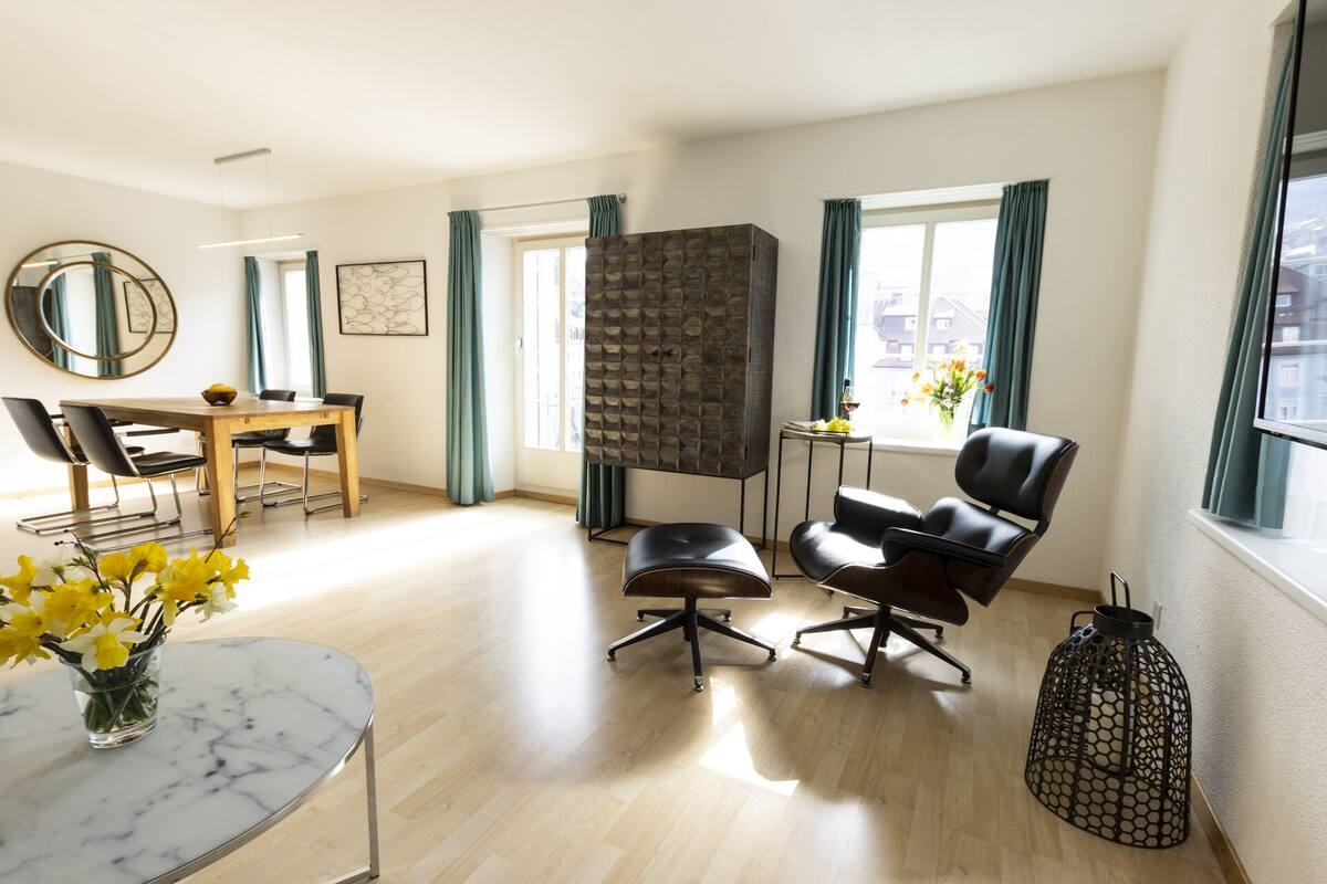 A modern living room with a dining area, a black leather armchair, a round coffee table, and a decorative vase with flowers.