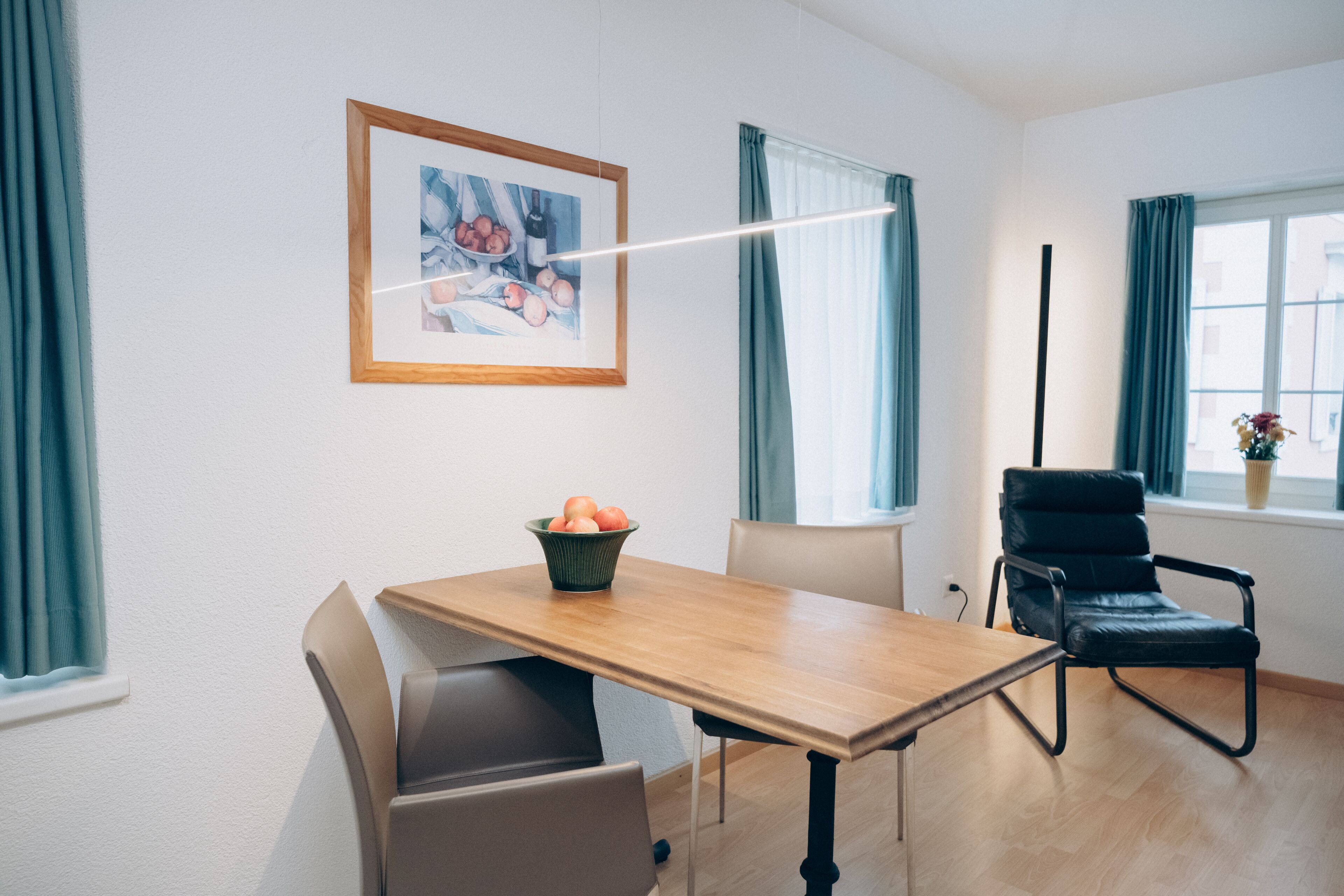 A modern room with a wooden dining table, a black leather chair, a framed artwork on the wall, and a window with curtains.
