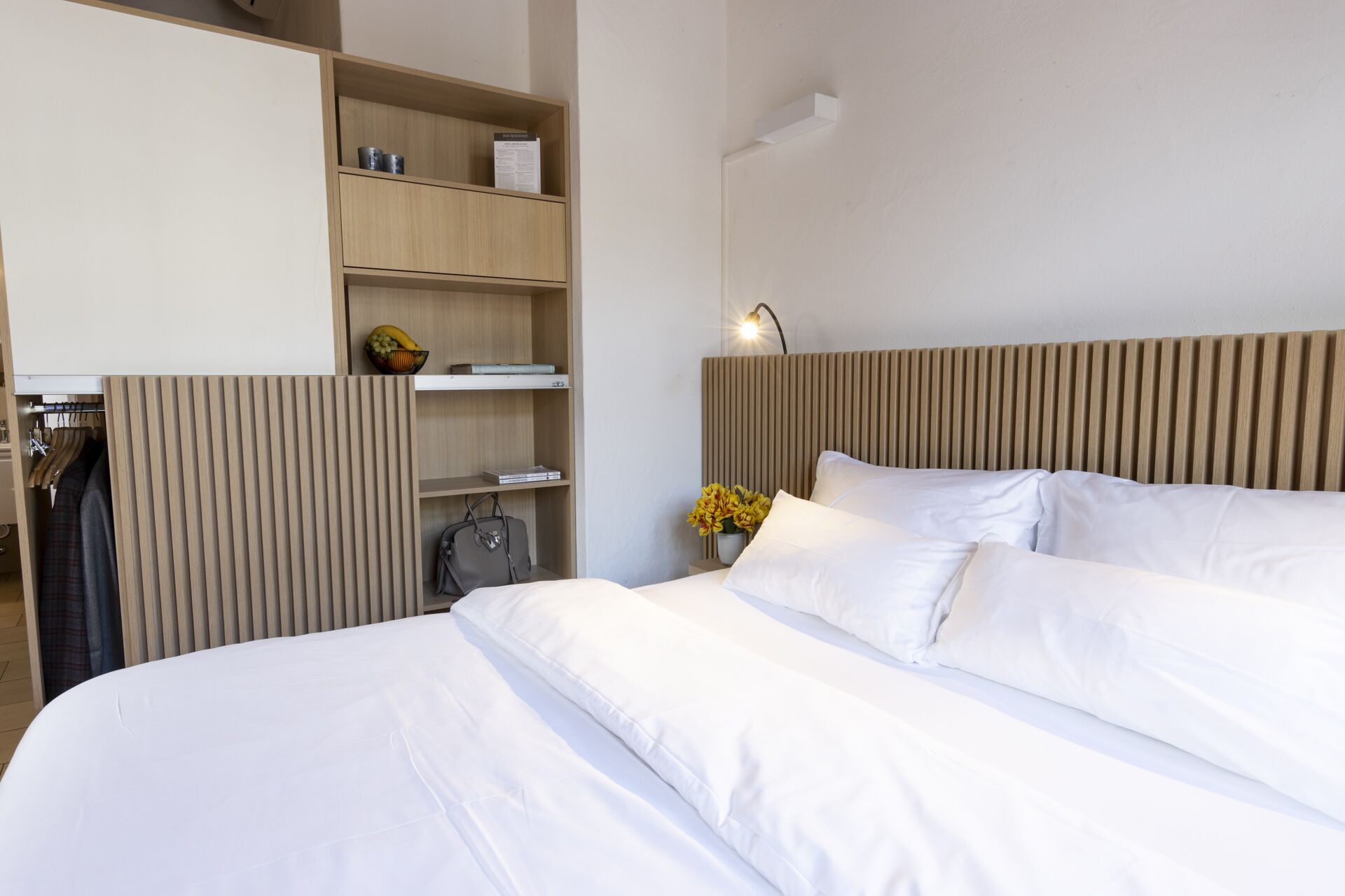 A hotel room with a bed, a wardrobe, a bedside table with a lamp, and a shelf with books and a fruit bowl.