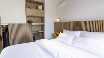 A hotel room with a bed, a wardrobe, a bedside table with a lamp, and a shelf with books and a fruit bowl.