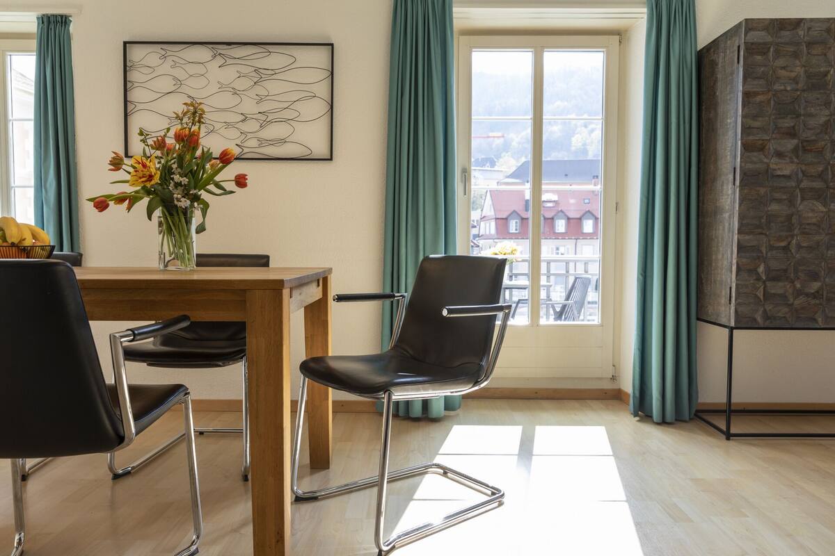 A modern room with a wooden dining table, two black chairs, a vase with flowers, a framed abstract art piece, a view of buildings through a window, and a stone-textured fireplace.