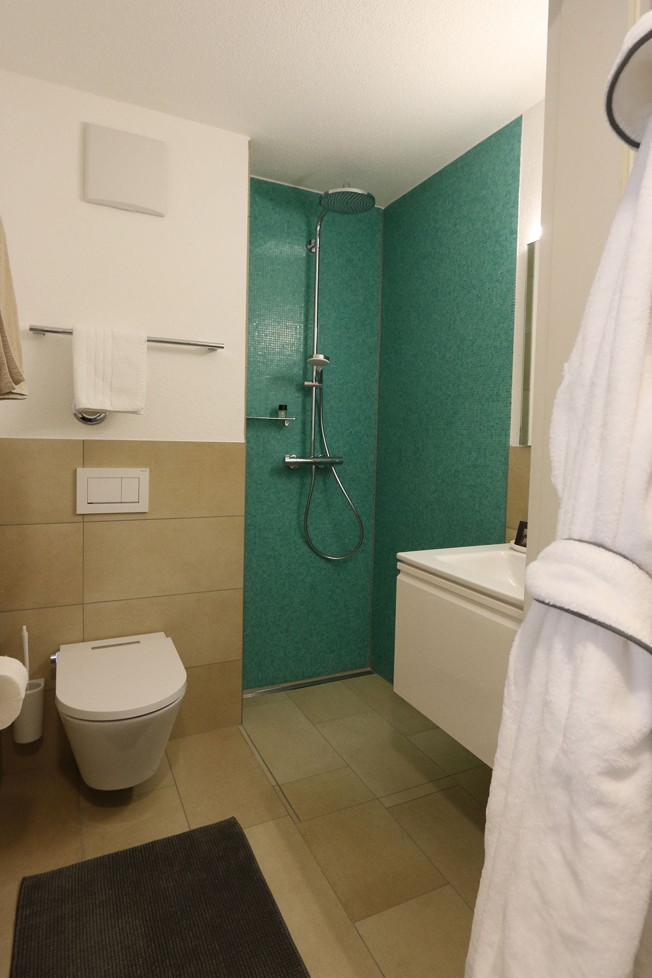 A bathroom with a toilet, bidet, and shower. There is a towel rack and a white robe hanging.