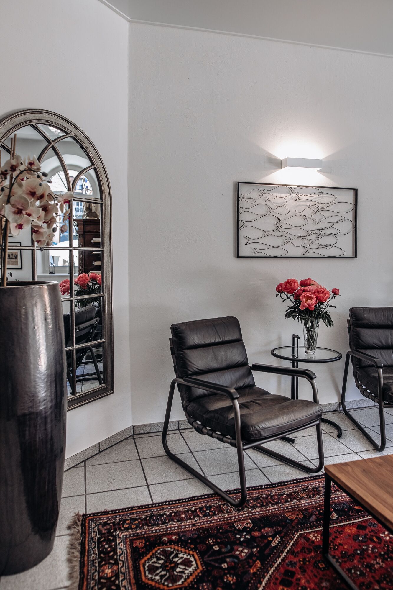A modern living room with a large mirror, two black leather chairs, a small round table with a vase of red flowers, and a patterned rug.
