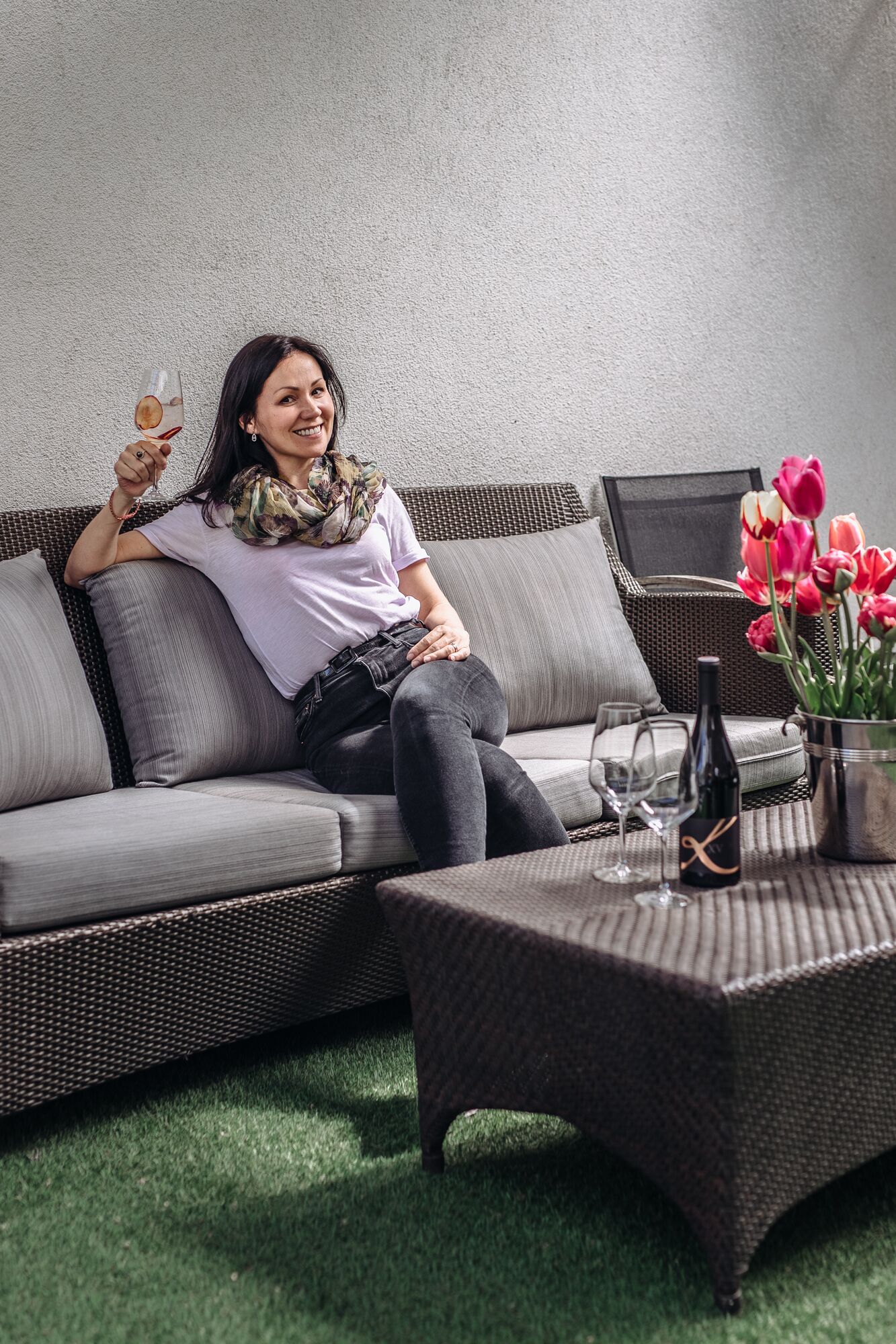 A person sitting on a grey sofa with a woven texture, a wicker coffee table with a bottle and glasses, and a vase with pink flowers.