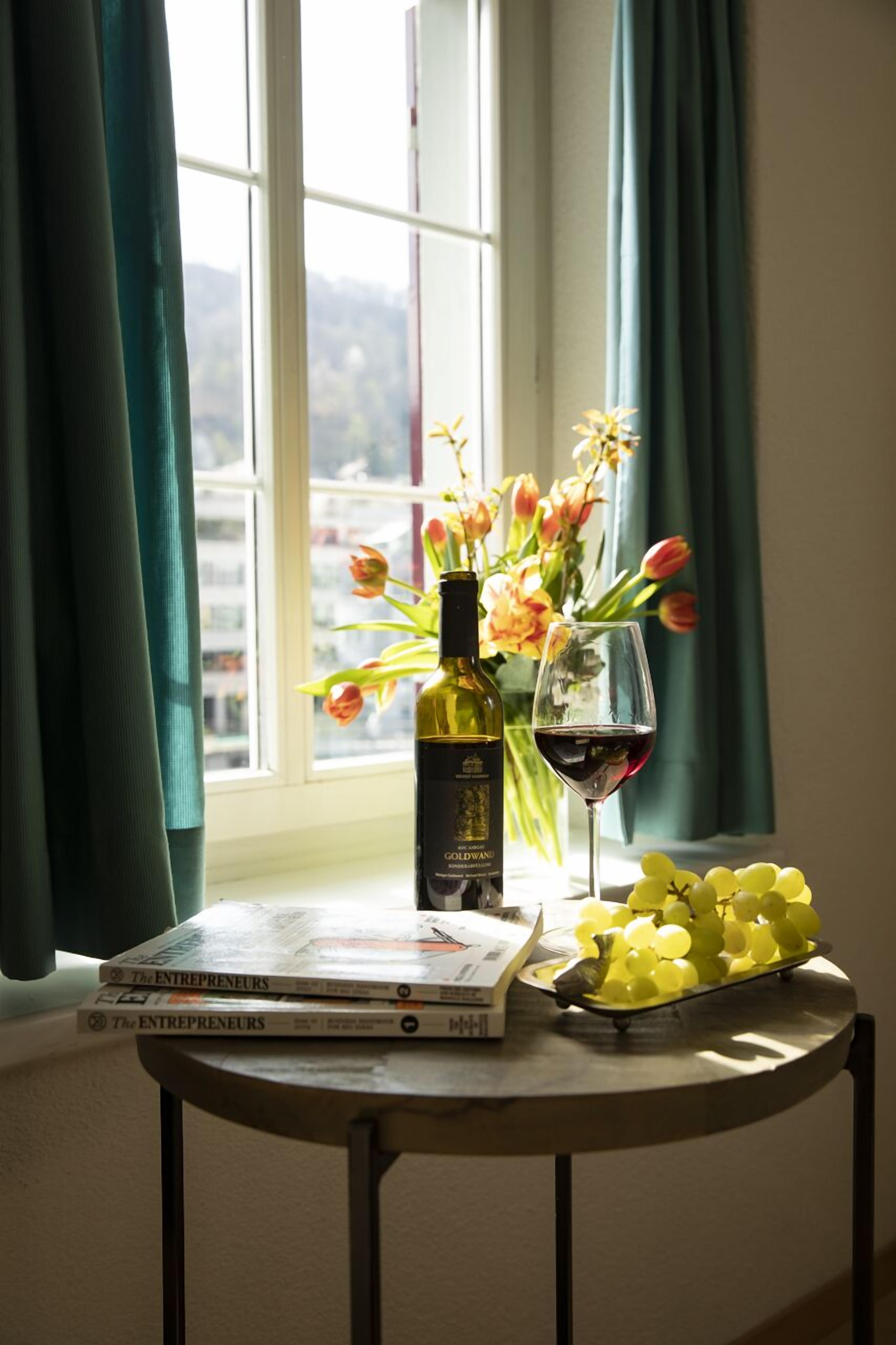 Ein Tisch mit einer Weinflasche, einem Weinglas, einem Blumenstrauß und einer Platte mit Trauben neben einem Fenster mit Vorhängen.