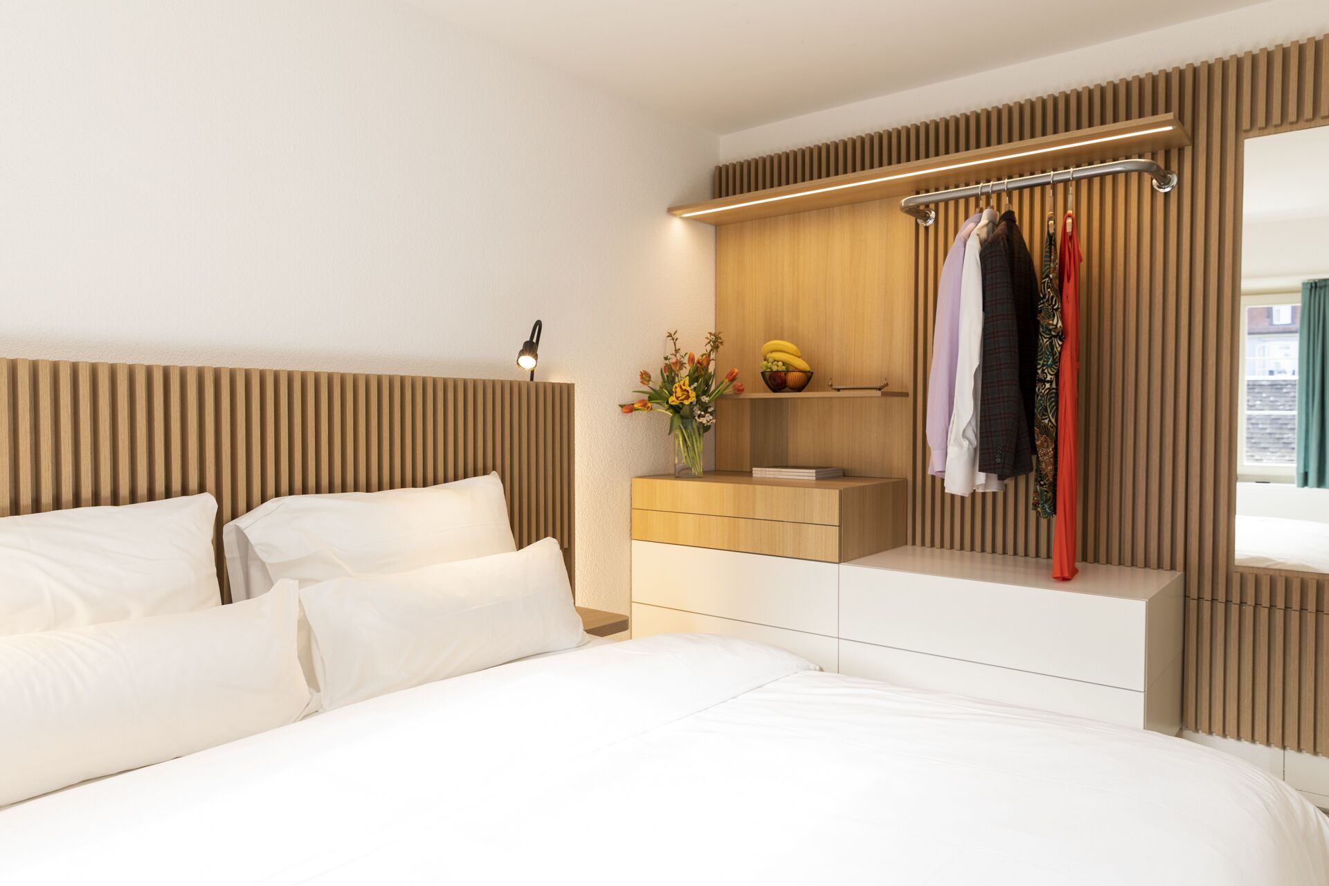 Une chambre moderne avec une tête de lit en bois, une table de chevet avec une lampe, une petite étagère ornée d’un vase de fleurs et un portant à vêtements.