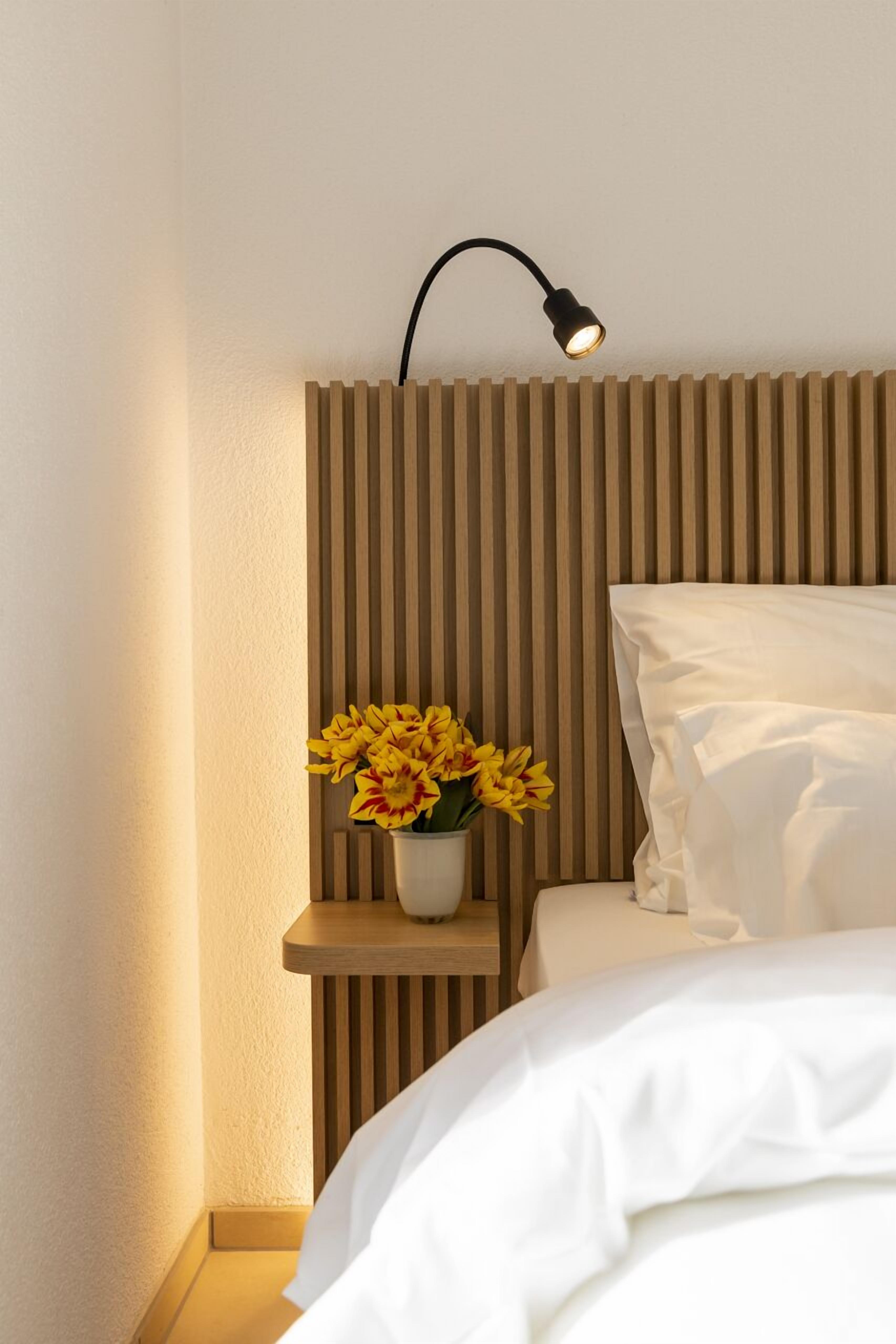 En pænt redt seng med hvide sengetøj, et natbord med en hvid vase med gule blomster, og en vægmonteret lampe.