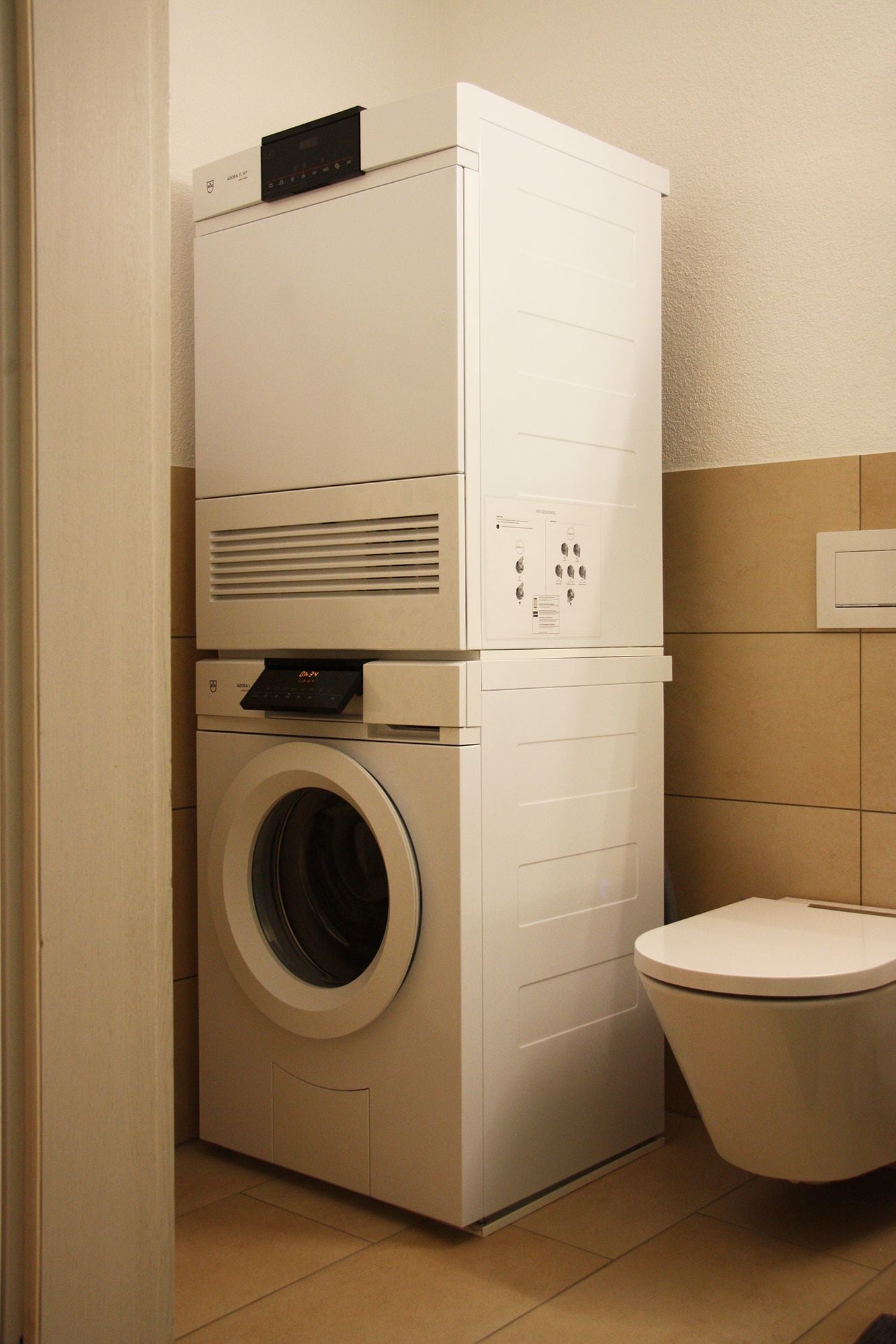 Un lavadero pequeño con lavadora y secadora de carga frontal, un inodoro y un enchufe eléctrico en la pared.