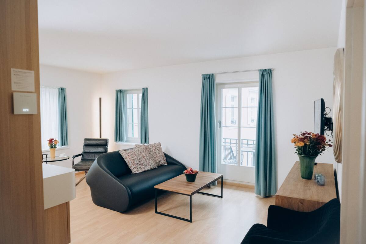 A modern living room with a black sofa, a wooden coffee table, and a dining table with a vase of flowers.