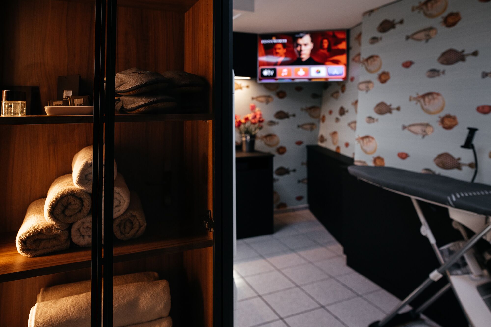 A room with a wooden shelf holding towels, a television on the wall displaying a sports channel, and a patterned wallpaper with fish.