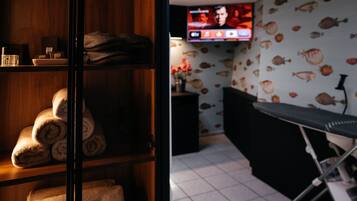 A room with a wooden shelf holding towels, a television on the wall displaying a sports channel, and a patterned wallpaper with fish.