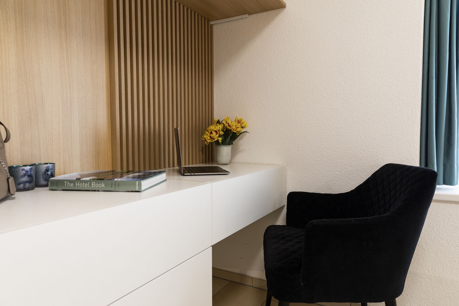 Una habitación moderna con un escritorio blanco, una silla negra, una computadora portátil, un libro titulado 'The Hotel Book', un florero con flores amarillas y un revestimiento de pared de listones de madera.