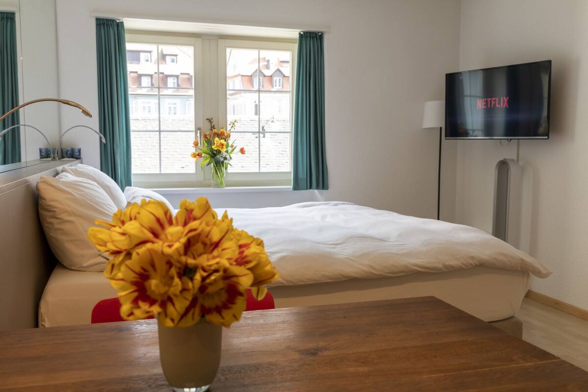 A hotel room with a bed, a TV on the wall, a window with curtains, and a vase with flowers on the bedside table.
