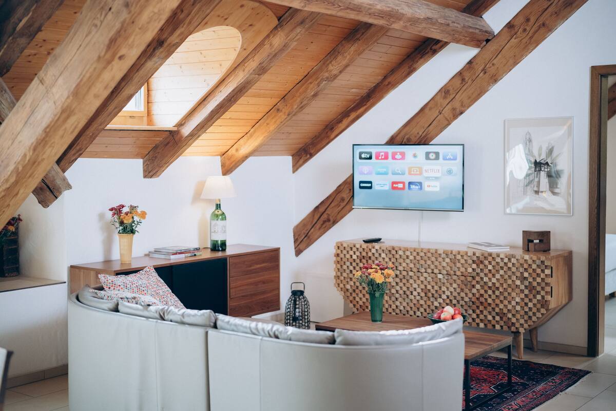 A modern loft with exposed wooden beams, a cozy seating area, a flat-screen TV, and a decorative wall piece.