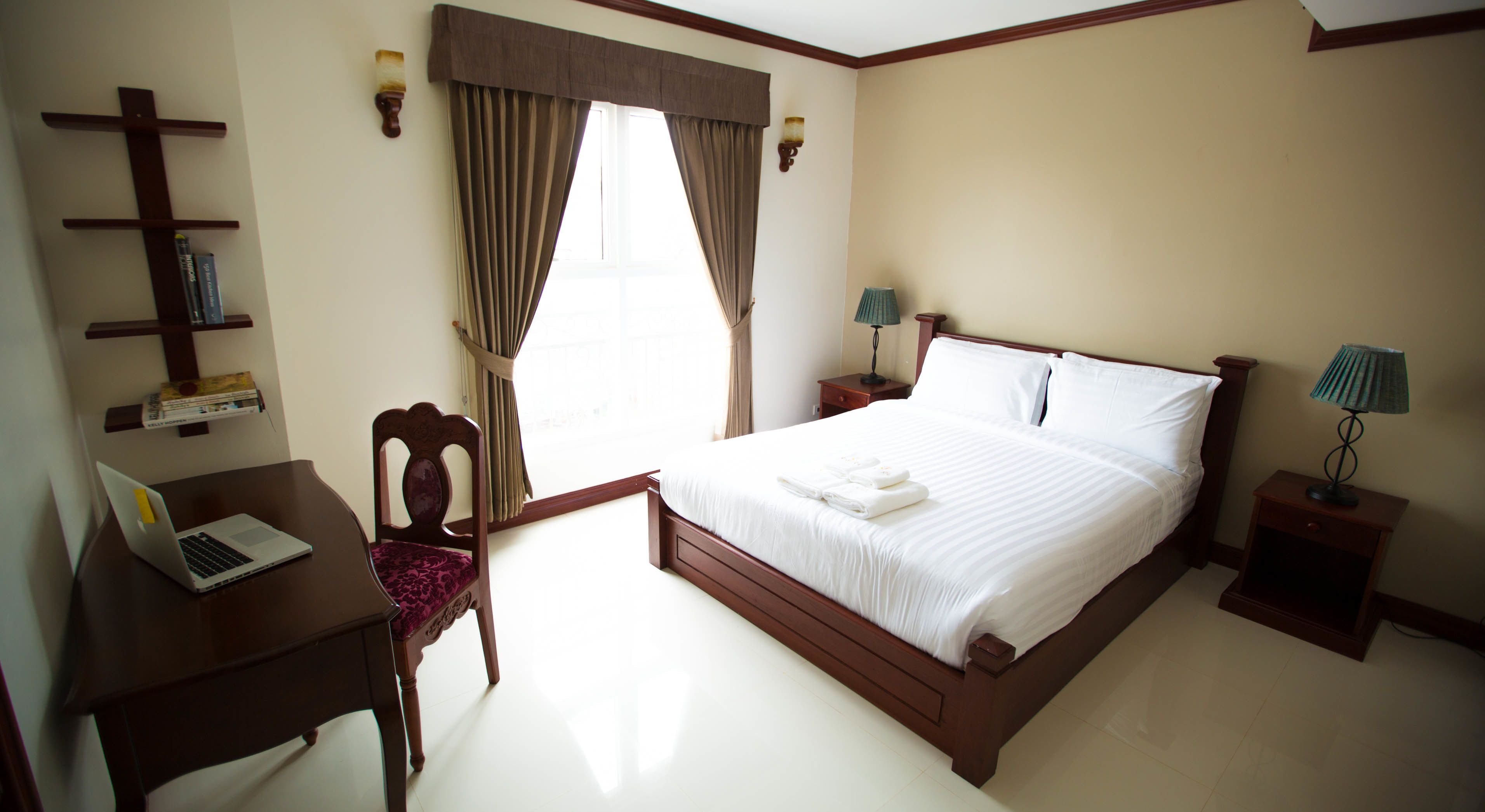 A hotel room with a wooden bed, a desk with a laptop, a chair, a nightstand with a lamp, and a window with curtains.