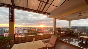 Un área de estar al aire libre techada con mesa y sillas, vista al atardecer sobre un paisaje urbano, y una barandilla.