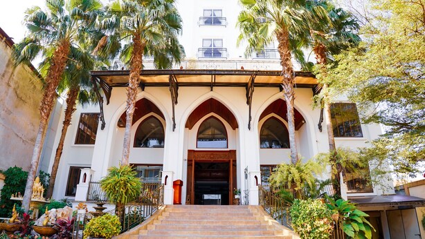 Un edificio con una entrada imponente, que presenta una escalera que conduce a una gran puerta de madera, flanqueada por dos altas palmeras.