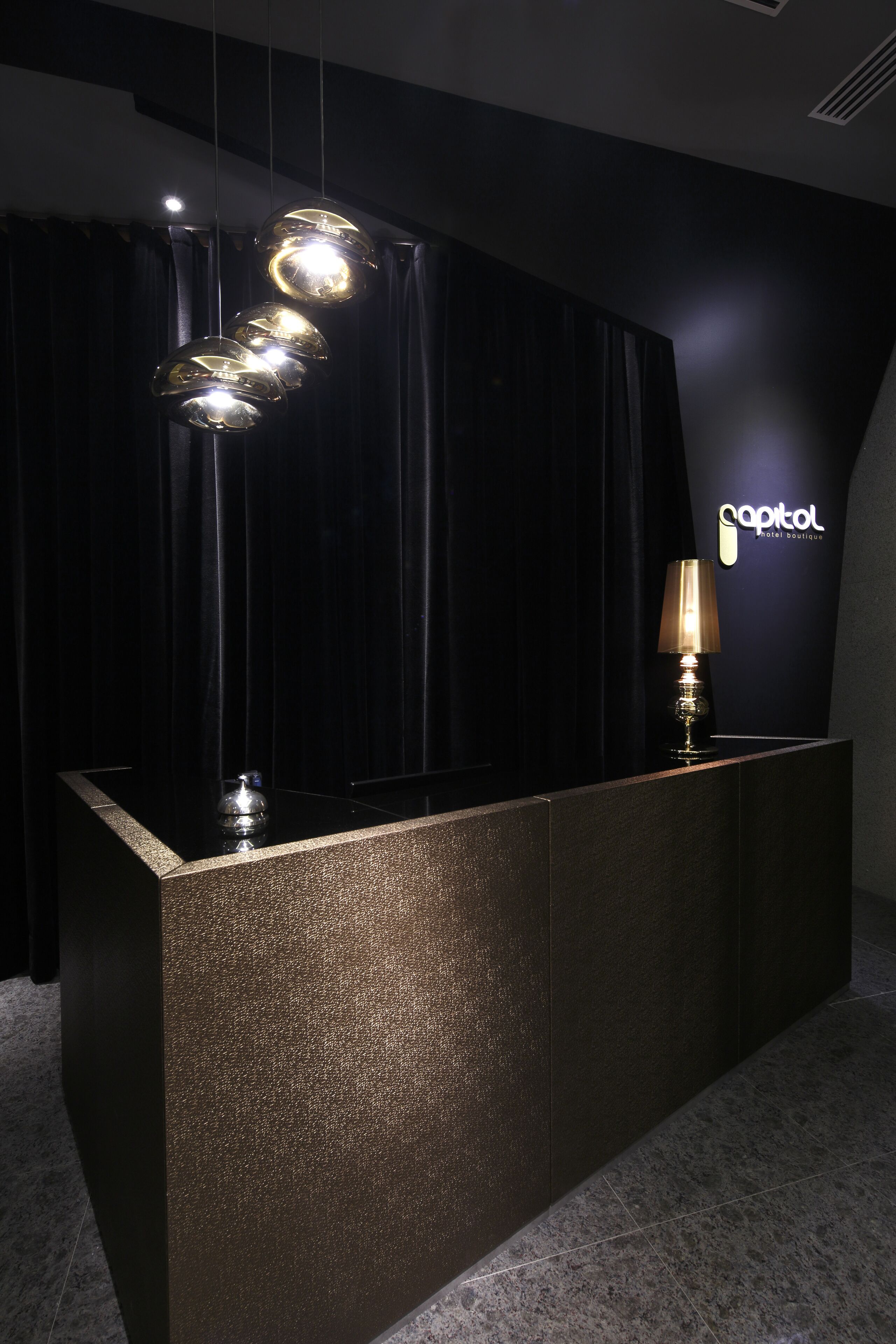 Un taulell de recepció amb una superfície fosca i amb textura, una làmpada i llums penjants. La paret del fons presenta un rètol amb el nom 'Capitol Hotel Boutique'.