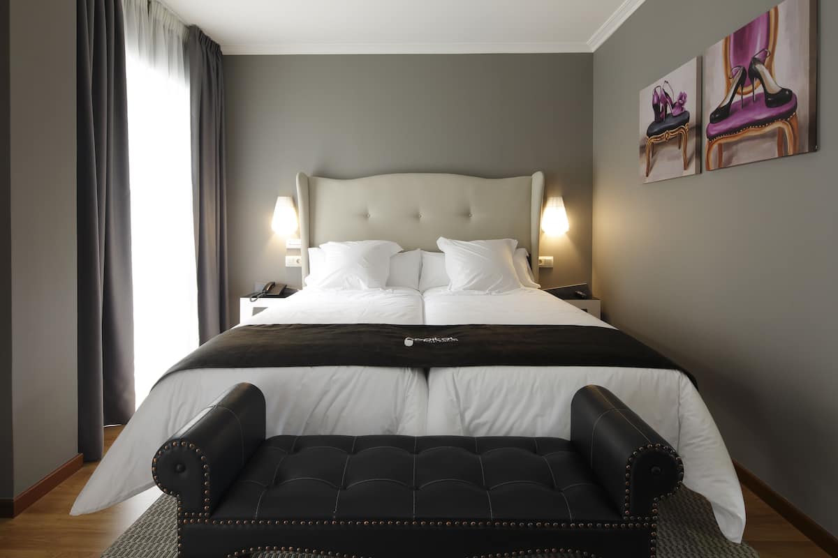 Une chambre d’hôtel avec un grand lit, deux tables de chevet, un banc noir recouvert de tissu et deux tableaux accrochés au mur.