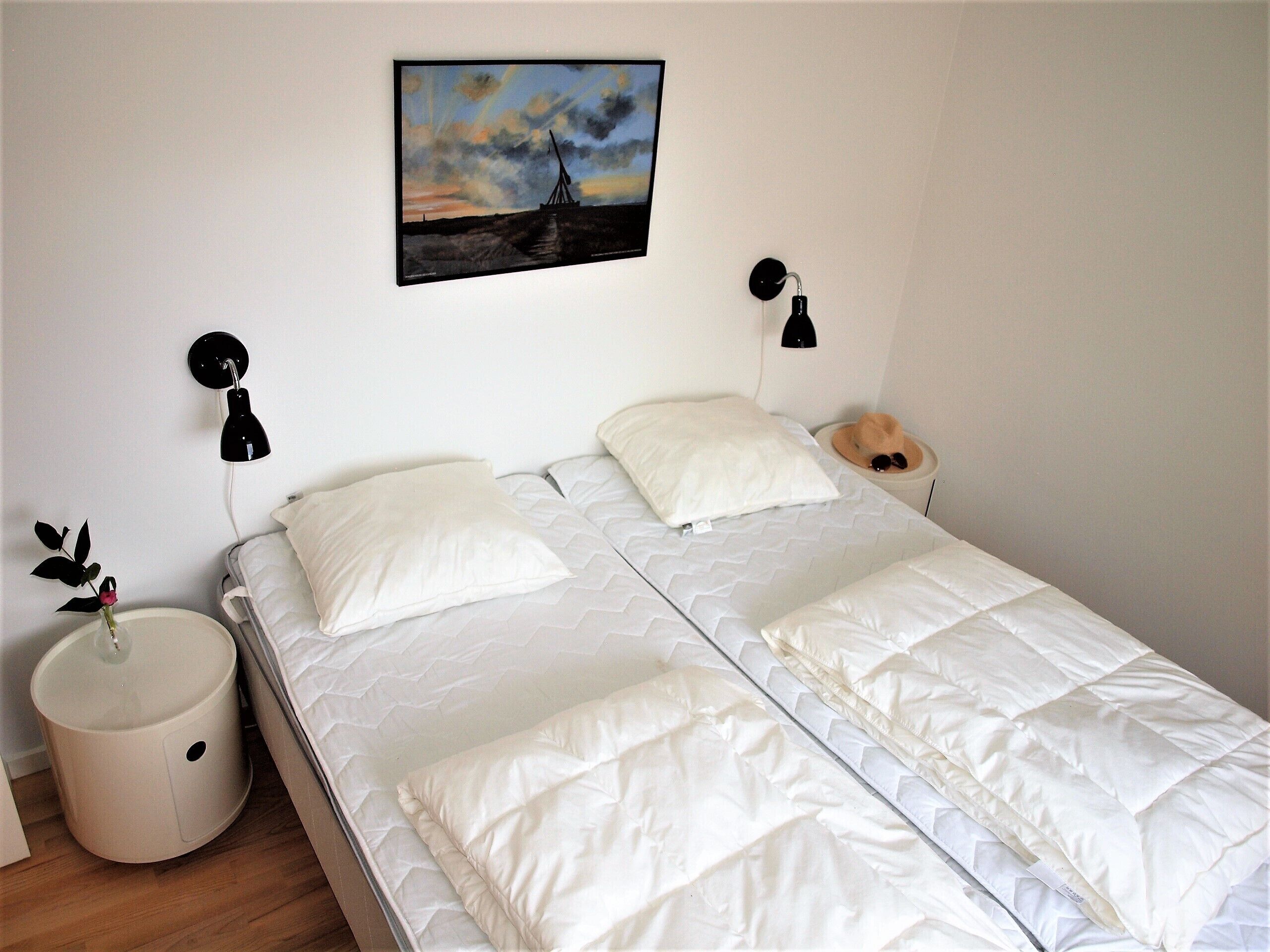 A neatly made bed with white bedding and two pillows, a bedside lamp, a small round nightstand with a vase of flowers, and a painting of a landscape on the wall.