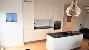 A modern kitchen with white cabinetry, a black countertop, and a built-in oven. There is a dining area with a table and chairs, and a black sofa. - Plesners Havnelejligheder (Skagen)