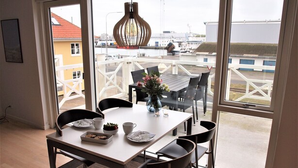 A dining area with a table set for four, a vase with flowers, and a view of the harbor through large windows. - Plesners Havnelejligheder (Skagen)
