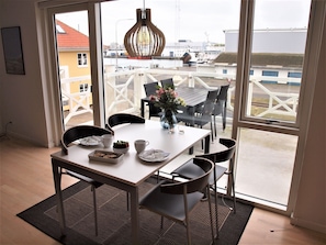 Un coin repas avec une table dressée pour quatre personnes, un vase rempli de fleurs et une vue sur le port, visible à travers de grandes fenêtres.