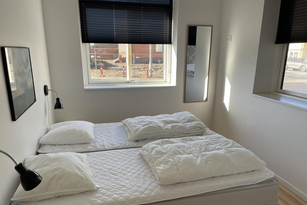A double bed with white bedding, a bedside lamp, a wall-mounted mirror, and a window with a view of construction outside.