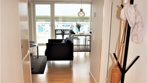A hallway leading to a dining area with a black sofa, a dining table, and chairs. There is a coat rack with a bag and a jacket on it. - Plesners Havnelejligheder (Skagen)