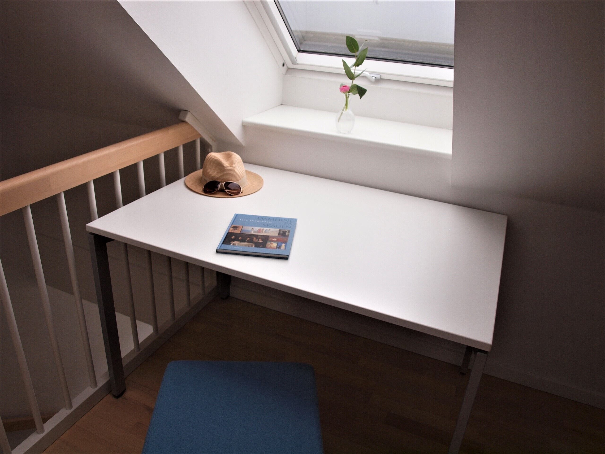 A small white table with a book and a hat on it, next to a window with a plant.