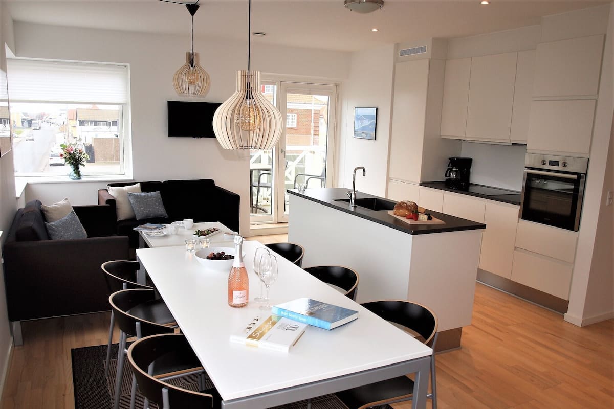 A modern kitchen with a central island, a dining area with a white table and chairs, and a living area with a sofa and a TV.
