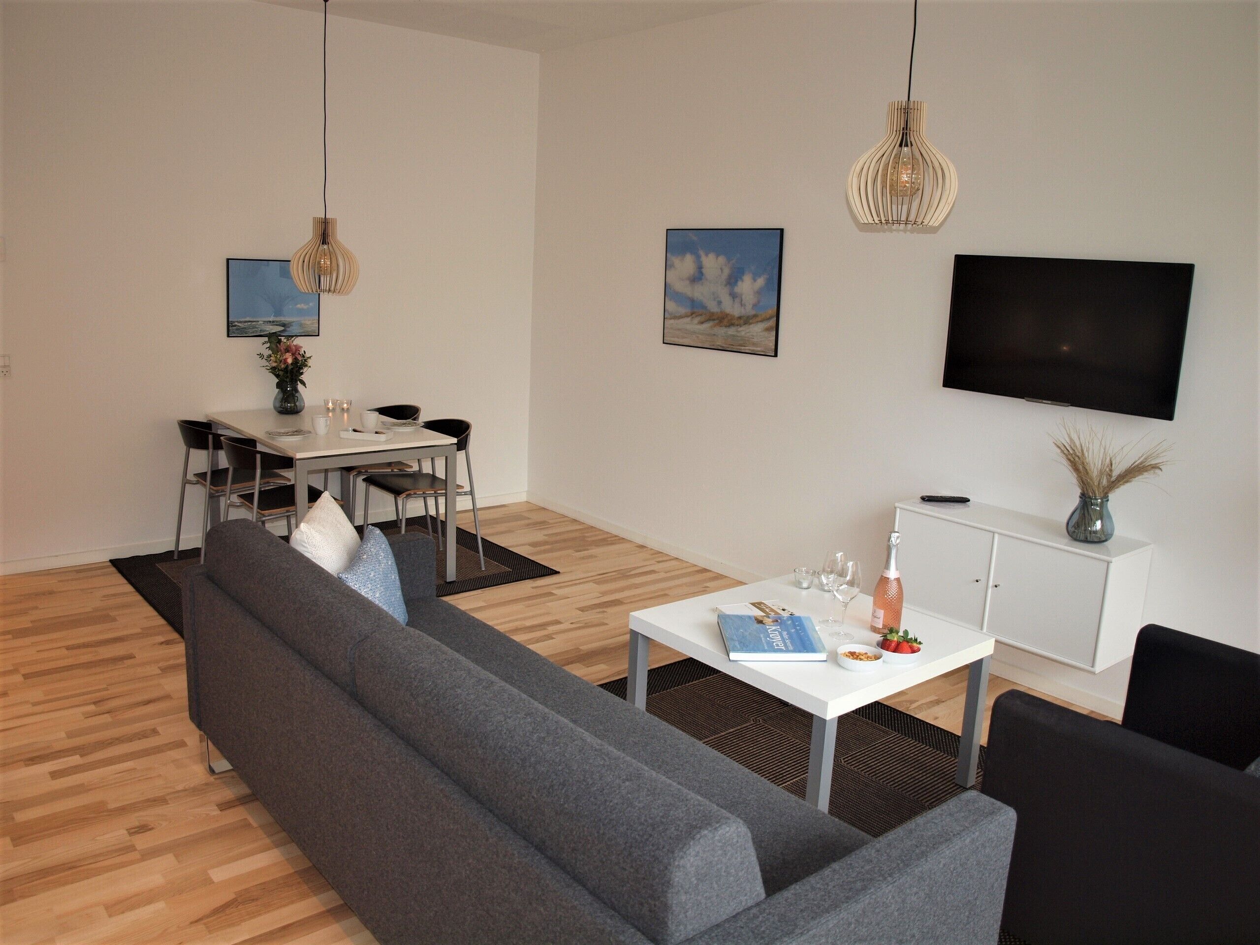 Un salon moderne avec un canapé gris, une table à manger et des chaises. Un téléviseur à écran plat est fixé au mur, et deux lampes suspendues sont placées au-dessus de la table.