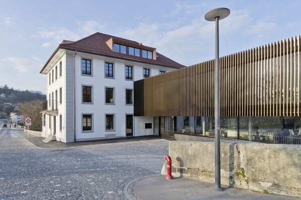 Un bâtiment moderne à la façade blanche et au toit en pente, situé à côté d’une structure en bois aux panneaux verticaux.