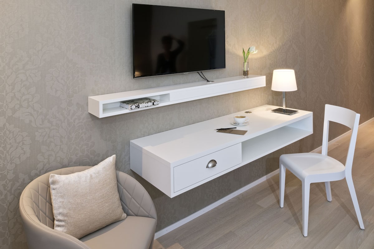 A modern living room with a flat-screen TV mounted on the wall, a white console table with a lamp, a white chair, and a sofa with a cushion.