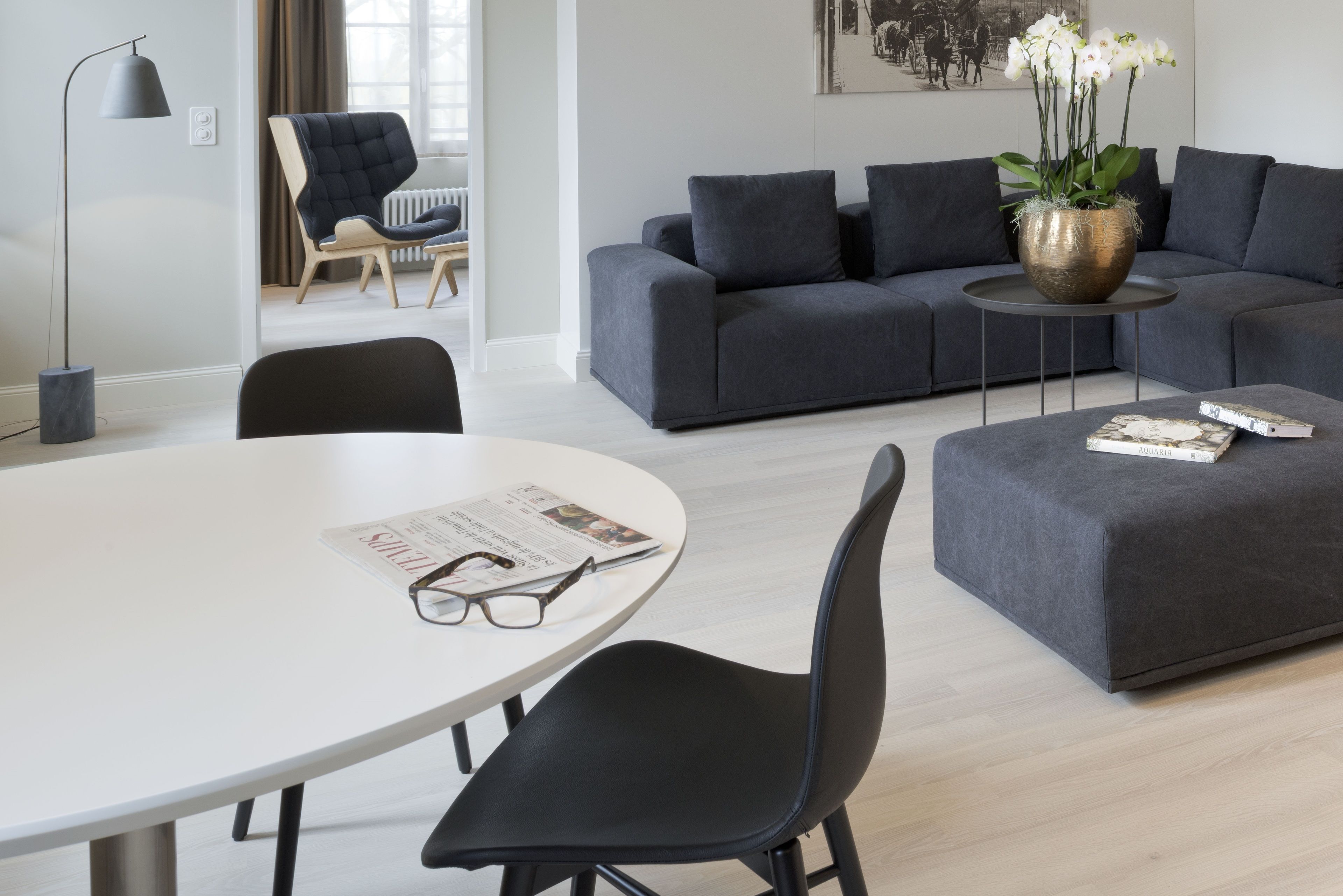 A modern living room with a grey sofa, a round coffee table, and a white dining table with chairs. There is a floor lamp, a potted plant, and a framed picture on the wall.